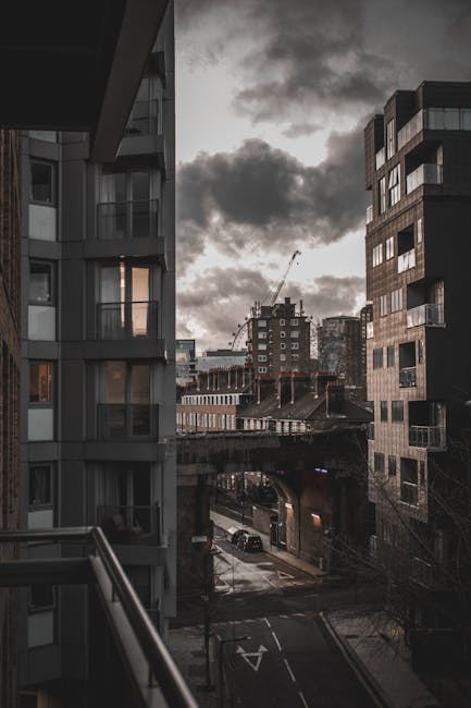 View from a balcony overlooking a narrow urban street in Bermondsey during late afternoon or early evening, with dark, cloudy skies overhead. The image shows two modern multi-storey residential buildi